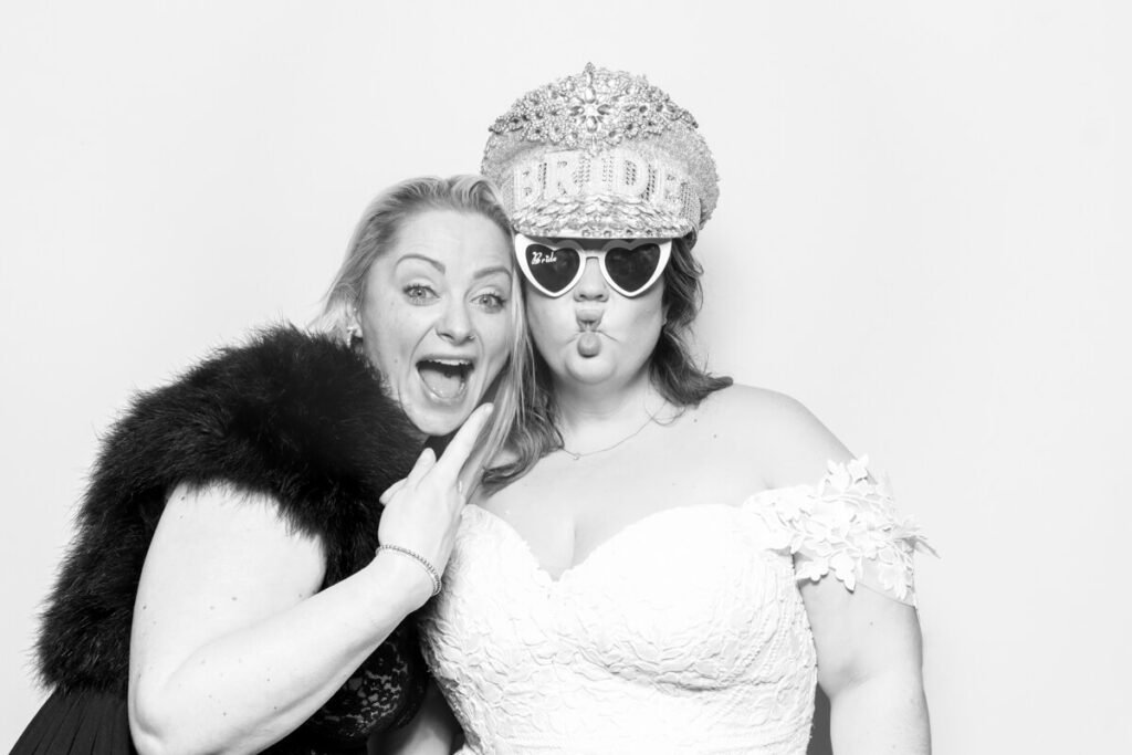 A bride and her friend posing in a black and white wedding photo booth. The bride wears a bejewelled 'Bride' hat and heart-shaped sunglasses, making a playful expression, while her friend smiles excitedly.