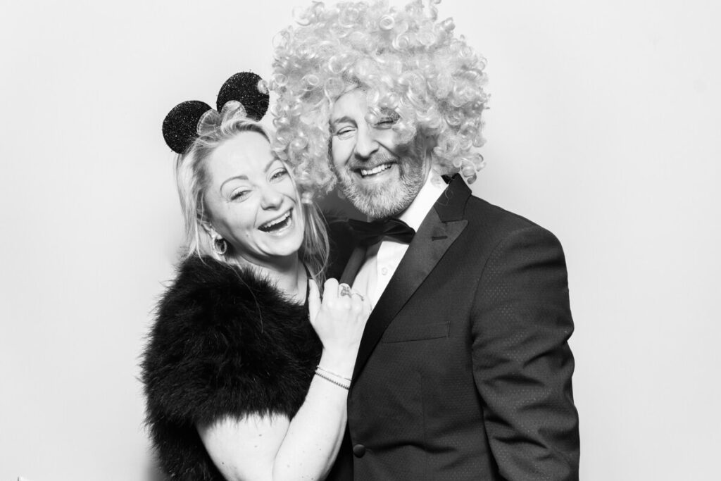  couple posing in a black and white wedding photo booth, laughing and dressed in formal attire. The woman wears Minnie Mouse ears, and the man sports a curly wig, adding a fun and playful touch to the moment