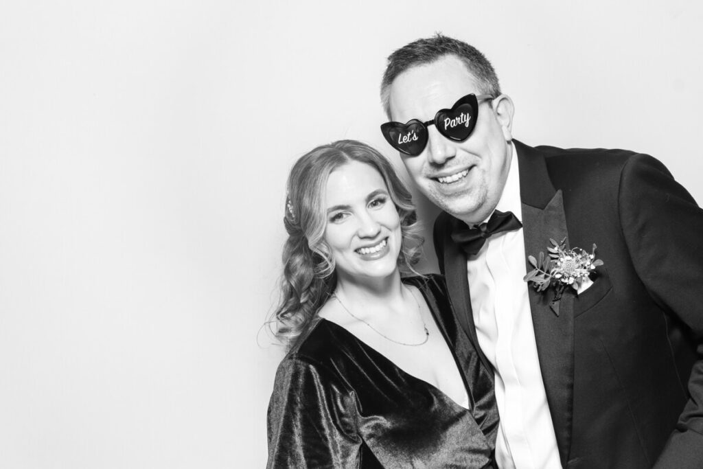 A couple posing in a black and white wedding photo booth. The woman is elegantly dressed in a velvet gown, while the man wears a tuxedo with a boutonnière and fun heart-shaped sunglasses that say 'Let's Party,' adding a playful touch to the moment