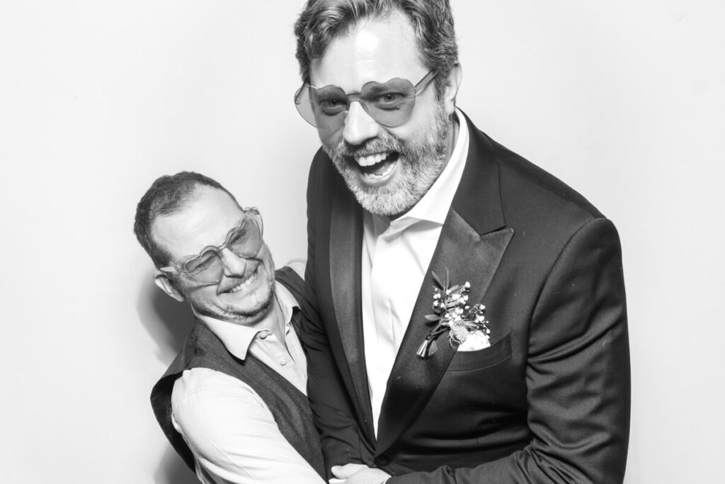 Two men dressed in formal wedding attire, laughing and posing playfully for the party entertainment. Both wear heart-shaped sunglasses, capturing a fun and joyful wedding moment.