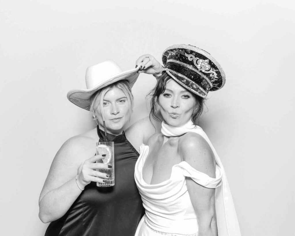 Two women posing in a black and white kardashian style booth, playfully dressed in a cowboy hat and a sequined captain’s hat. One holds a drink while the other pouts, capturing a fun and stylish wedding moment.