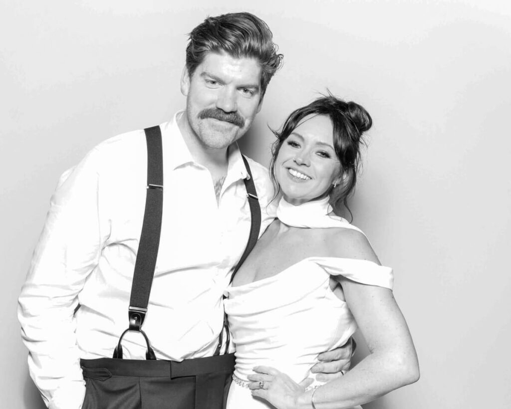 A couple posing in a black and white wedding photo booth, dressed in elegant attire. The groom wears suspenders, and the bride wears an off-shoulder dress, both smiling warmly for a timeless and stylish photo