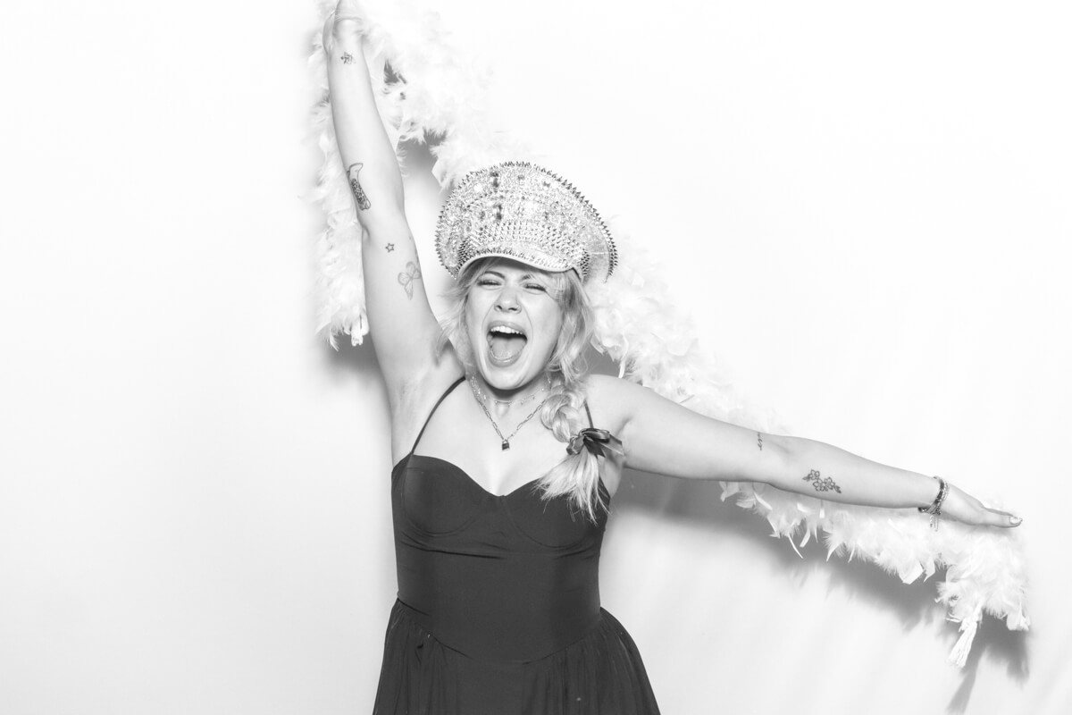 A joyful woman in a black and white wedding photo booth, wearing a sparkling embellished hat and a feather boa, posing energetically with her arms raised and a wide smile.