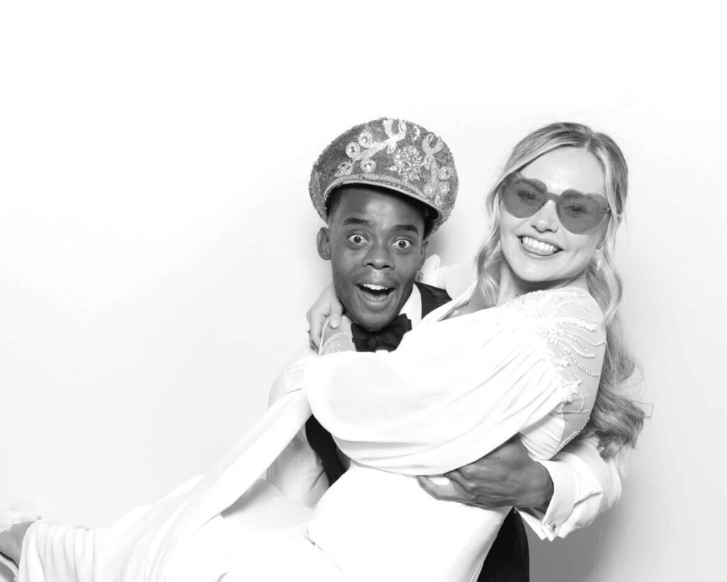 A joyful wedding couple posing in a black and white photo booth, with the groom playfully lifting the bride. Both are dressed in formal attire, wearing fun accessories, and sharing a lighthearted moment