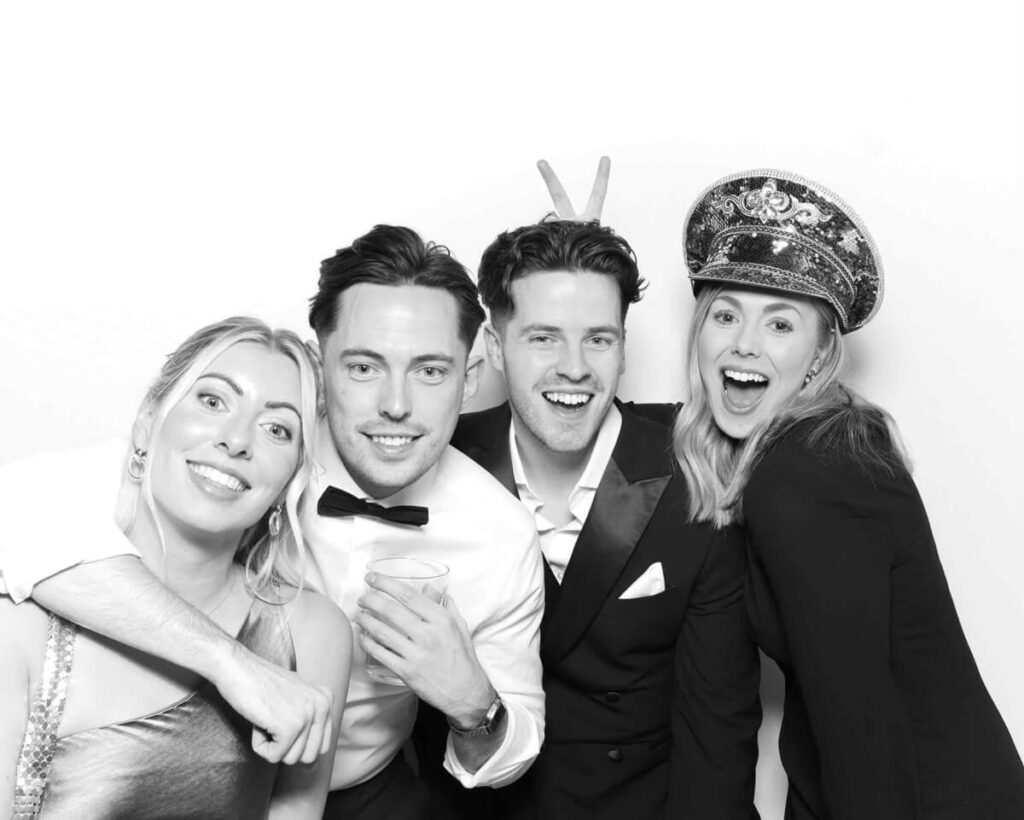 A group of four wedding guests posing in a black and white photo booth, dressed in formal attire with playful expressions and props, capturing a fun and stylish moment.