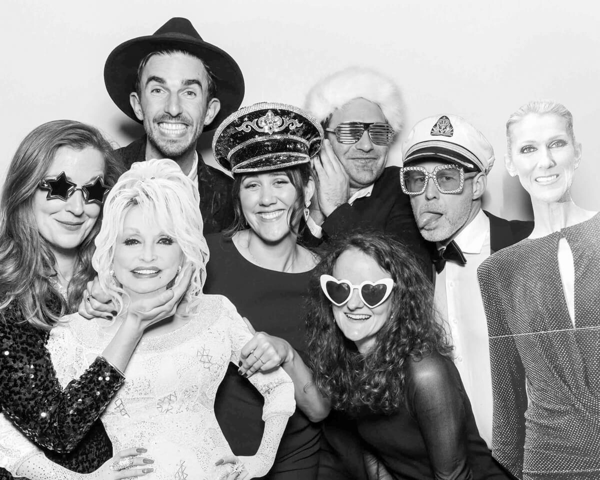 A group of people dressed in fun costumes and accessories, posing in a black and white wedding photo booth with celebrity cutouts and stylish props, creating a lively and entertaining atmosphere