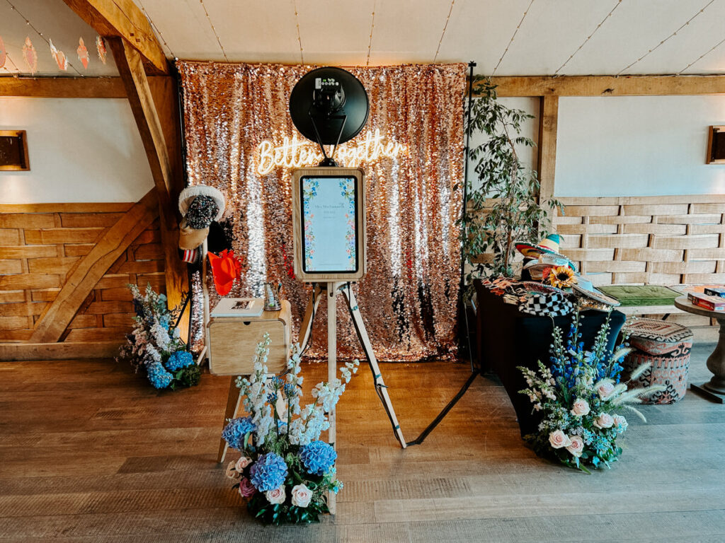 A stylish wedding photo booth setup in a rustic venue, featuring a rose gold sequin backdrop with 'Better Together' neon signage, a professional camera on a tripod, floral decorations, and a table with fun props for guests.