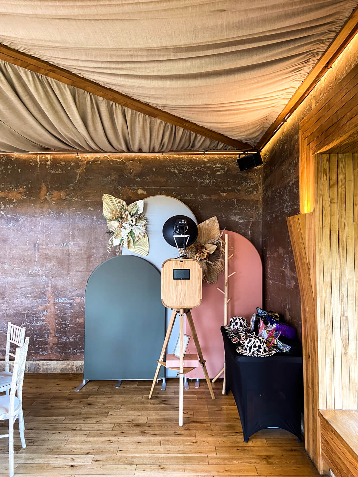 A stylish photo booth setup at Elmore Court in Gloucester, featuring a modern wooden photo booth on a tripod, pastel arched backdrops with floral accents, and a table filled with fun props for guests.