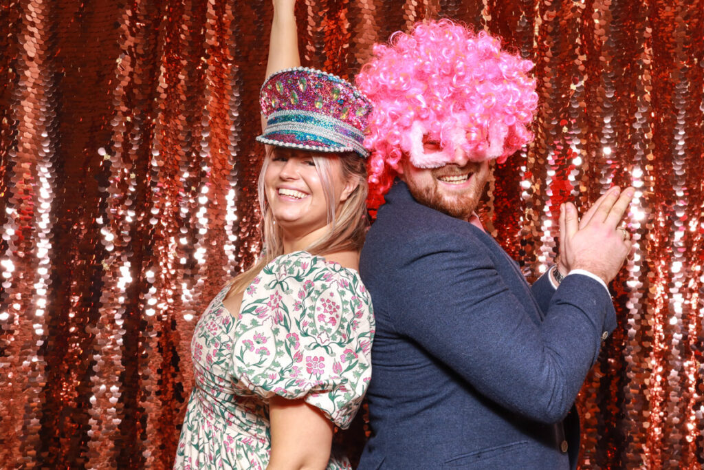 2 wedding guests having lots of fun wearing curated props and posing back to back against a champagne sequins backdrop, during a party entertainment service, by mad hat photo booth