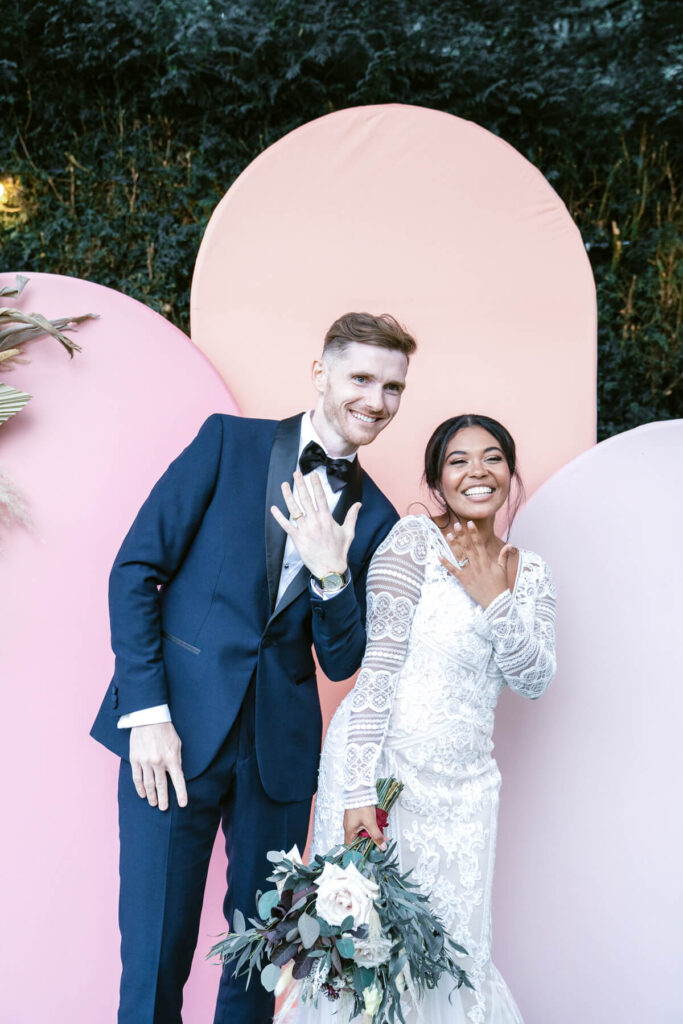 bride and groom posing with their new wedding rings on, against a modern arch photo booth backdrop, during the wedding party entertainment in the Cotswolds