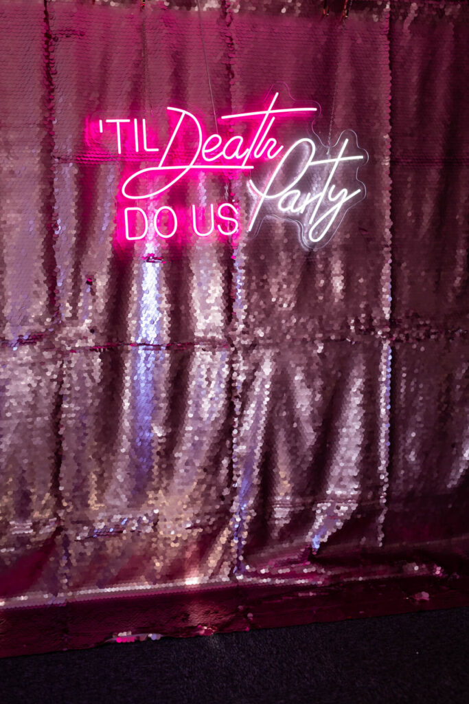 "Till Death Do Us Party" neon sign available for hire and rental , against a lilac champagne sequins backdrop , perfect backdrop for a wedding ceremony, reception and party 