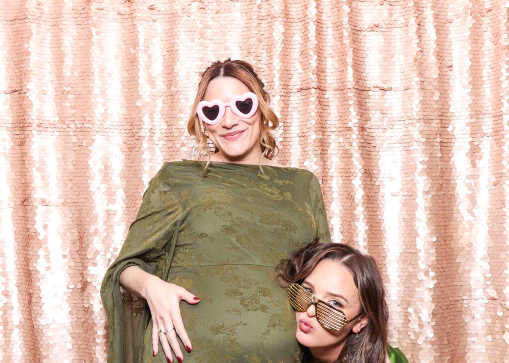 2 guests posing with fancy props, against a blush sequins backdrop, for a selfie pod private party hire in Cotswolds area