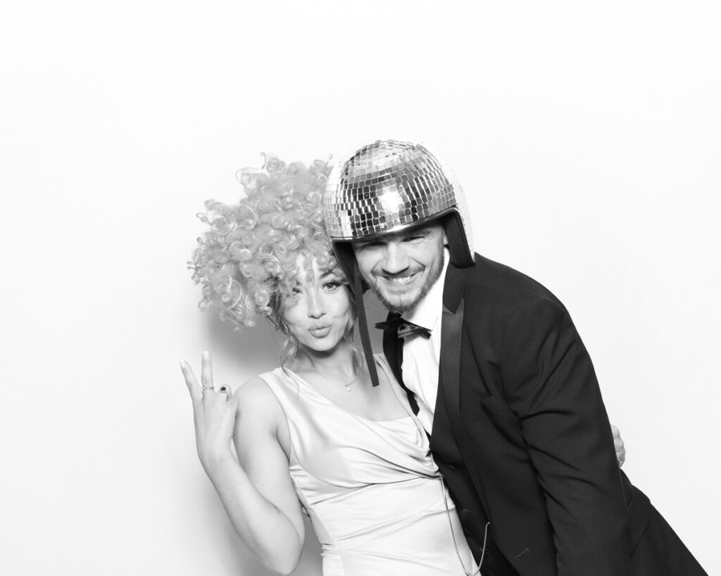 a black and white image of a couple wearing fun party props and disco ball hat, posing against a white backdrop, for a photo booth hire in the cotswolds