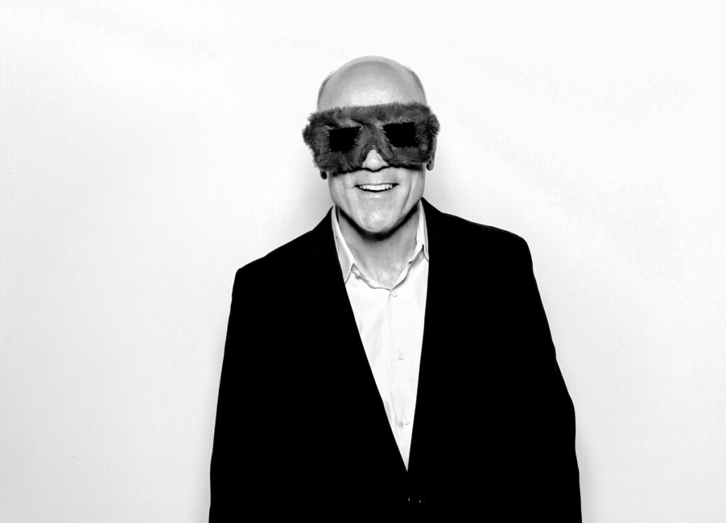 a black and white image of a man wearing furry party sunglasses, posing against a white backdrop, for a photo booth hire in the cotswolds