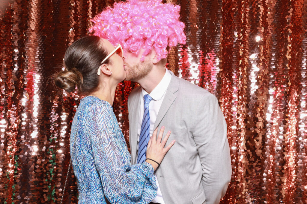 2 guests posing and kissing while wearing fancy props, against a champagne sequins backdrop, for a selfie pod private party hire in Cotswolds area