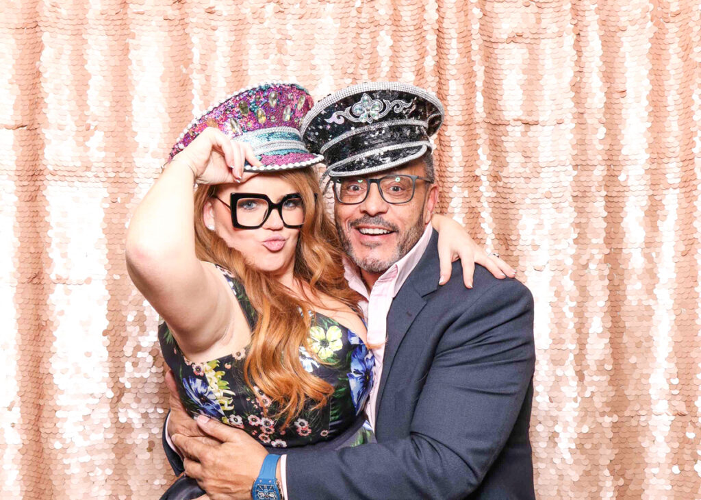 2 guests posing with fancy props, against a blush sequins backdrop, for a selfie pod private party hire in Cotswolds area