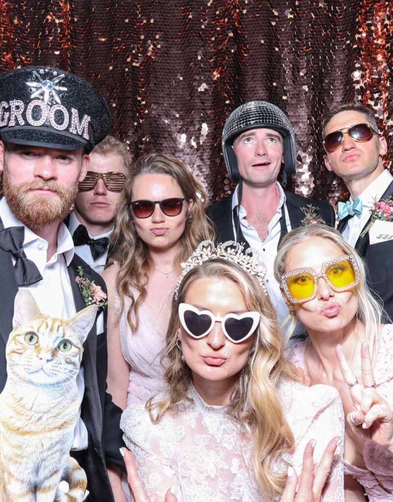 large group of guests posing with super fun props, against a champagne sequins backdrop, for a cripps barn cotswolds wedding party entertainment with mad hat photo booth hire for parties, birthdays and weddings