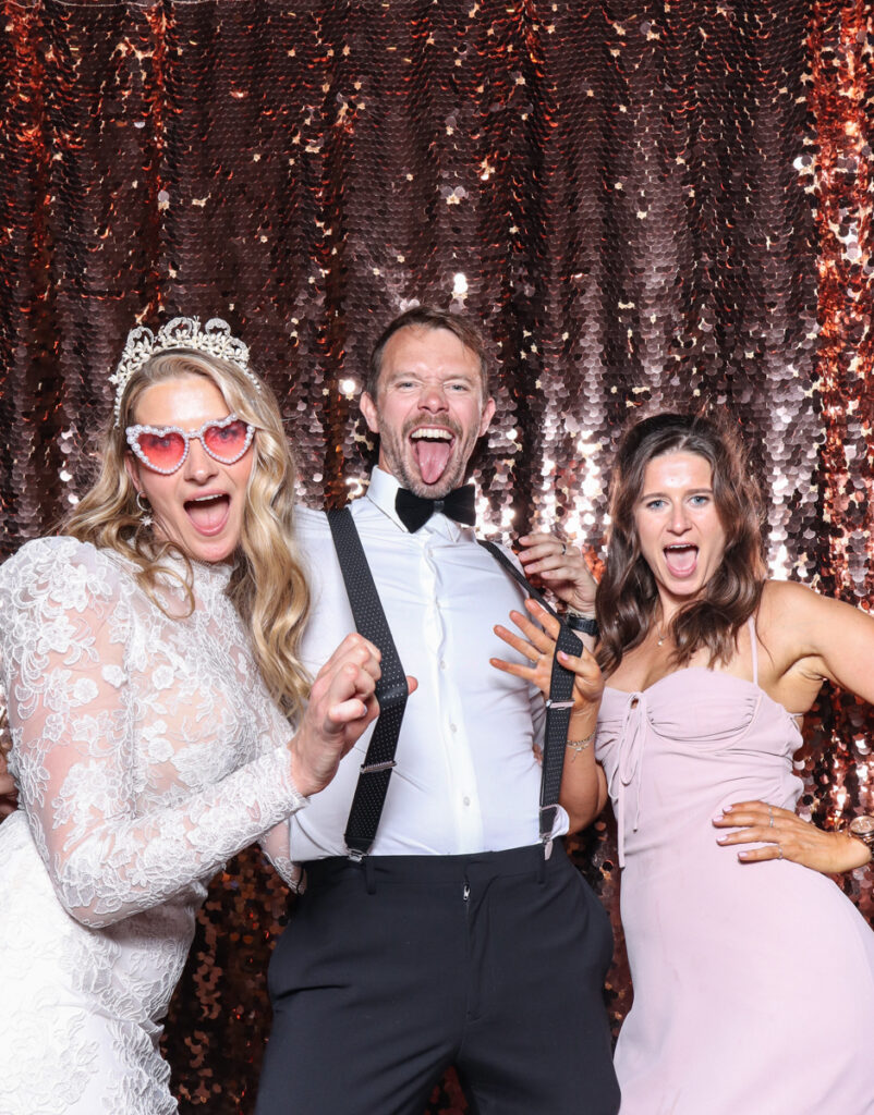 3 guests posing with super fun props, against a champagne sequins backdrop, for a cripps barn cotswolds wedding party entertainment with mad hat photo booth hire for parties, birthdays and weddings
