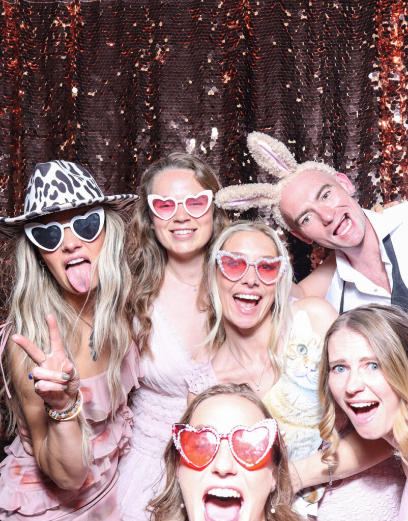 large group of guests posing with super fun props, against a champagne sequins backdrop, for a cripps barn cotswolds wedding party entertainment with mad hat photo booth hire for parties, birthdays and weddings