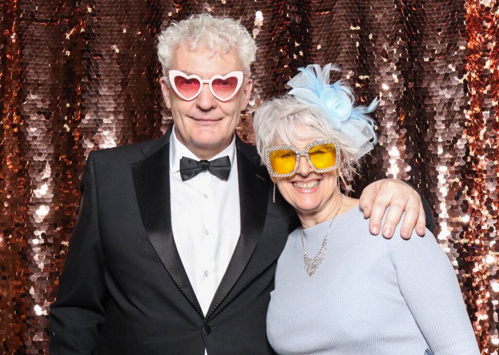 2 elderly guests posing with super fun props, against a champagne sequins backdrop, for a cripps barn cotswolds wedding party entertainment with mad hat photo booth hire for parties, birthdays and weddings