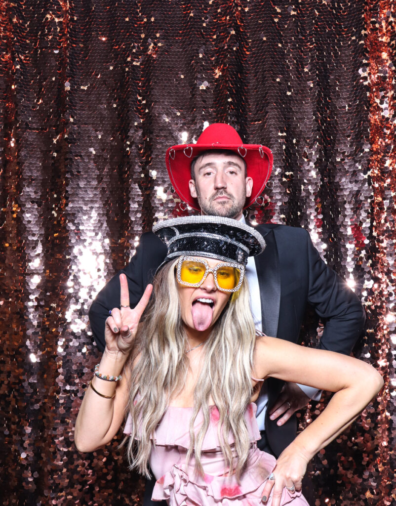 2 guests posing with super fun props, against a champagne sequins backdrop, for a cripps barn cotswolds wedding party entertainment with mad hat photo booth hire for parties, birthdays and weddings