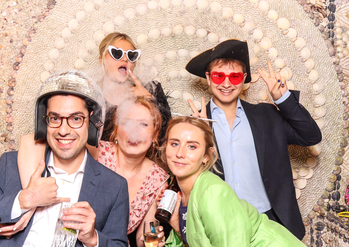 A group of 5 guests laughing and posing with props, drinks, and bright accessories against an ornate shell backdrop.