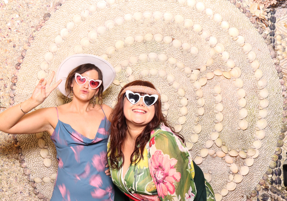 Two women posing playfully with heart-shaped sunglasses and hats in front of a detailed shell wall backdrop.