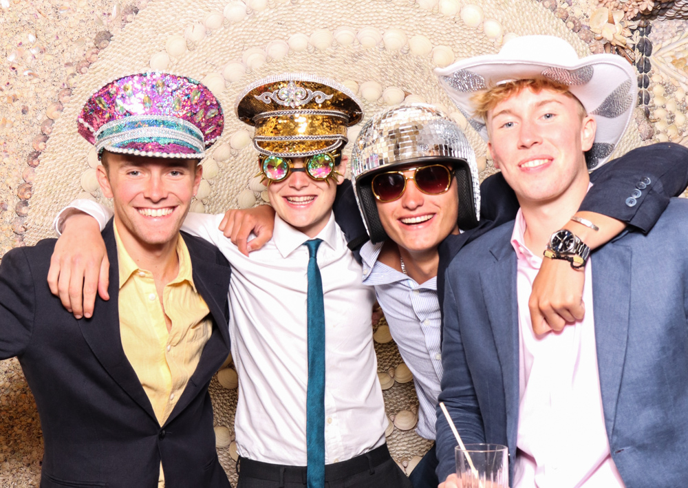Group of four young men in party hats and props, smiling together against an ornate spiral shell pattern.