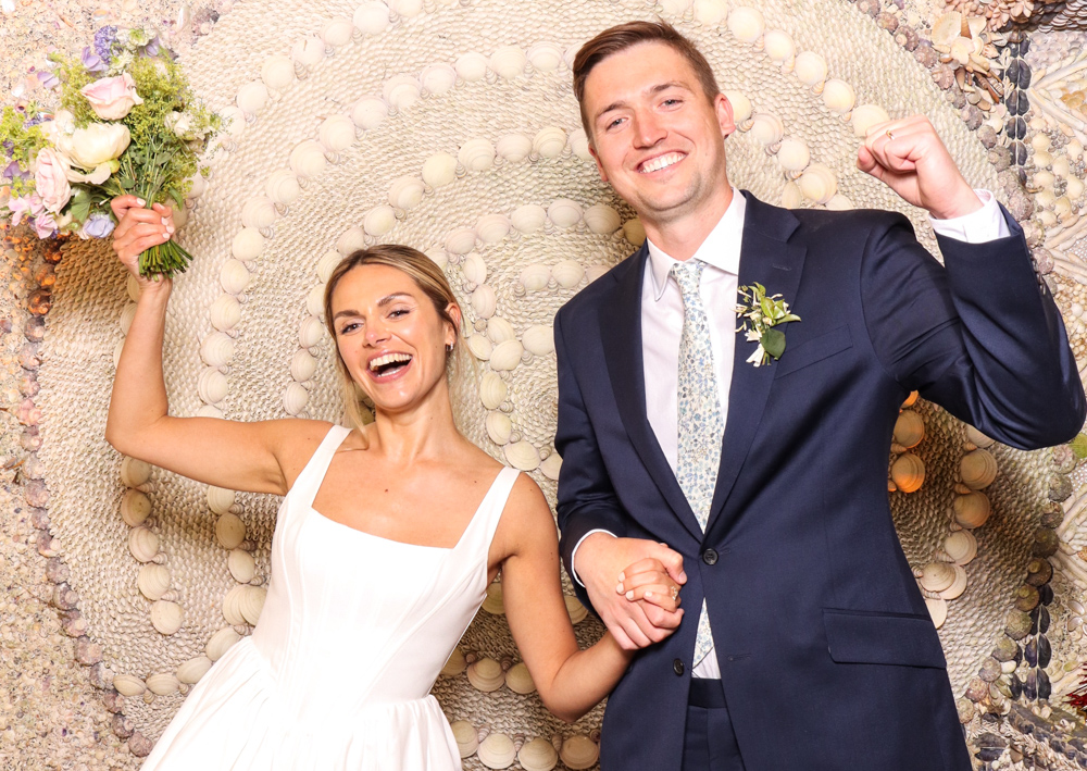 Bride and groom celebrating with a bouquet and big smiles, standing in front of a textured shell backdrop at Belcombe Court wedding venue