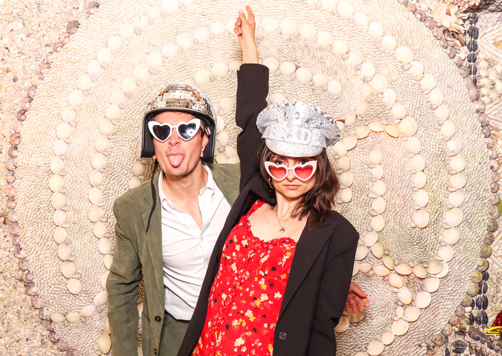 Couple striking a playful pose with heart-shaped glasses and bold props in front of a shell-patterned backdrop.