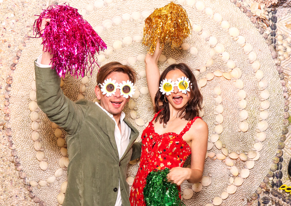 Two guests celebrating with colourful pom-poms and daisy-shaped glasses, mid-laugh in front of a spiral shell wall at Belcombe Court