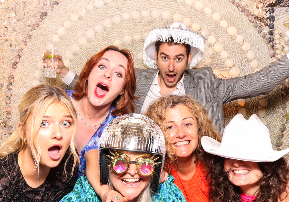 Group of six friends pulling fun faces and wearing an assortment of party hats and props, framed by a textured wall of shells.