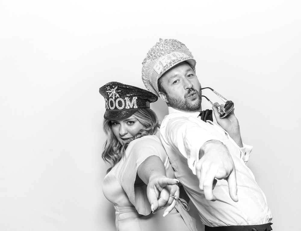 Bride and groom pointing towards the camera while wearing matching themed hats and fun props during their photo booth session.