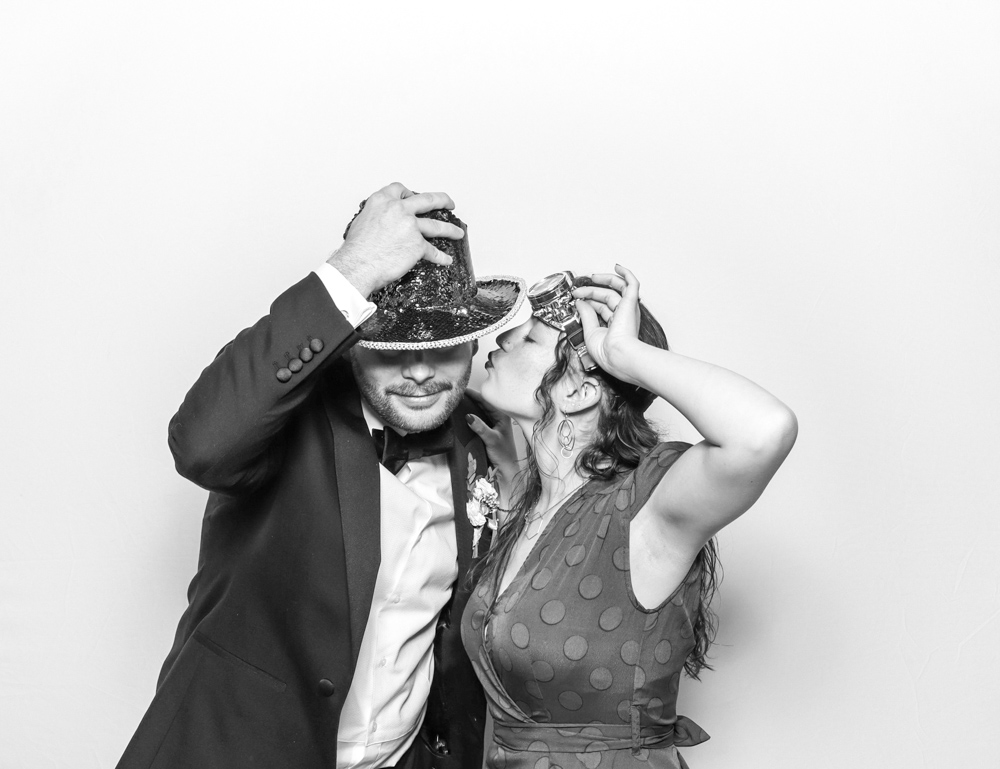 Couple dressed up in fun party hats, sharing a kiss while adjusting their props in the photo booth.