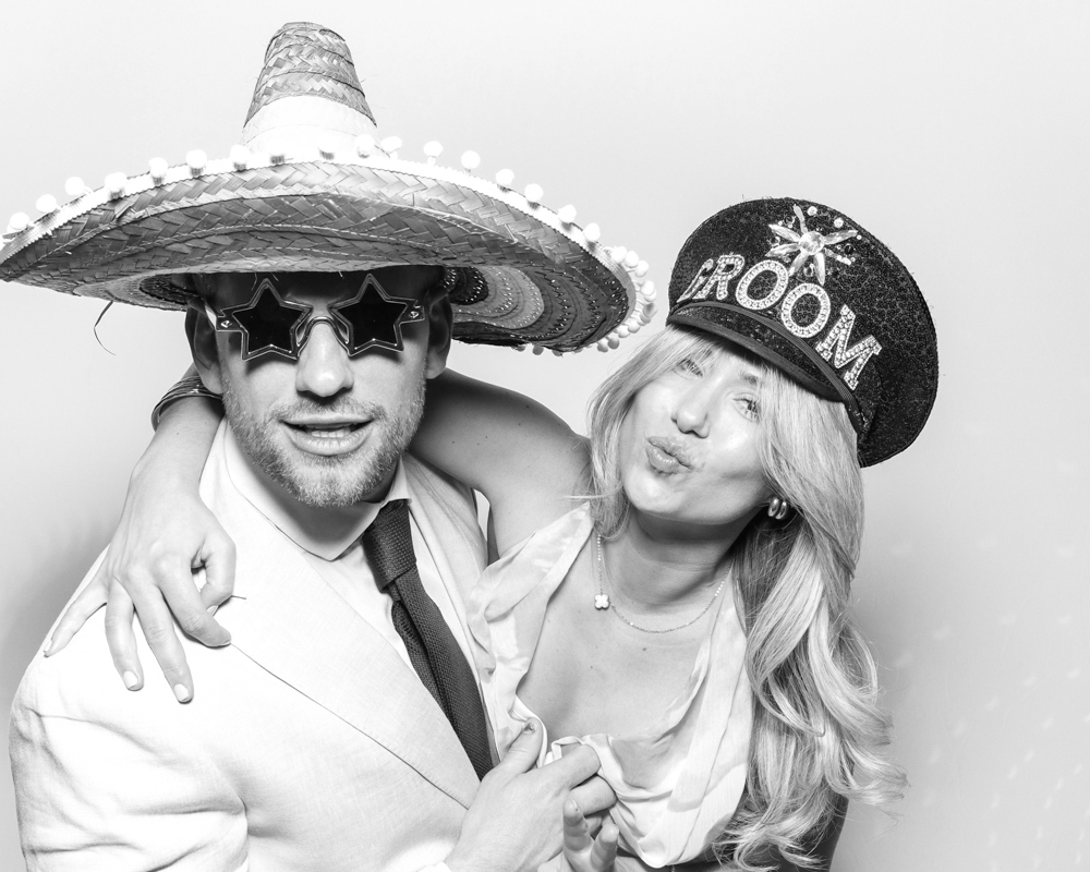 Man in a sombrero and star-shaped glasses posing with a woman wearing a “GROOM” hat, both making playful expressions.