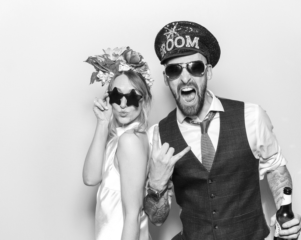 Energetic couple in themed outfits and sunglasses striking dramatic poses, including a “GROOM” hat and beer in hand.