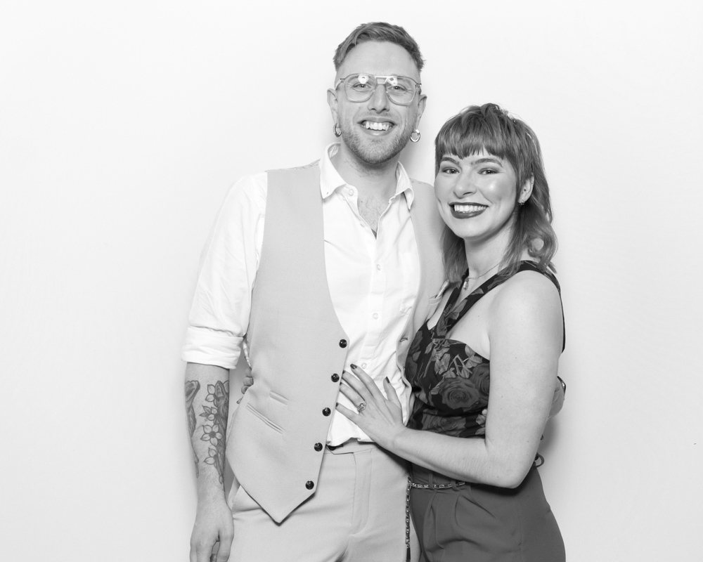 Smiling couple at an event posing in front of a white backdrop, captured by a digital photo booth with a black and white finish.