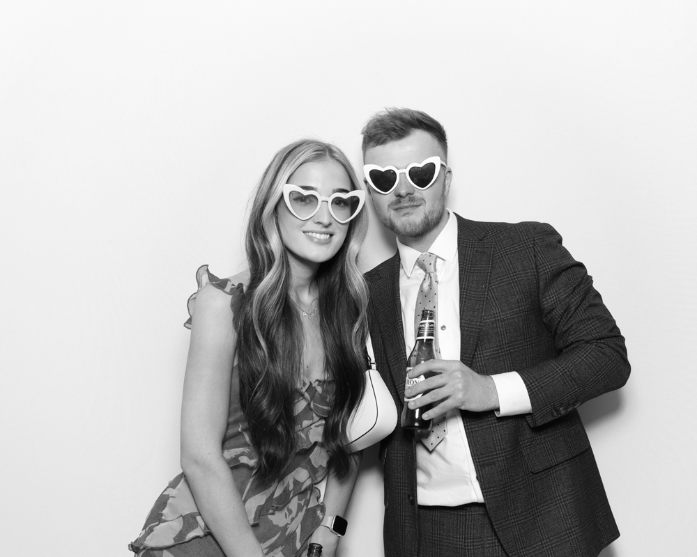 Couple wearing heart-shaped sunglasses holding drinks, taken in a digital photo booth with a monochrome filter.