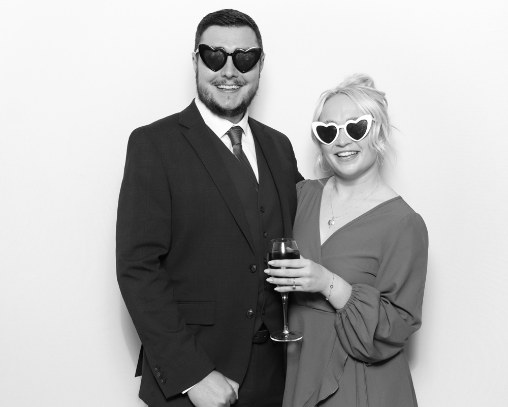 Couple wearing heart-shaped sunglasses and smiling in a digital photo booth with a glass of wine in hand.