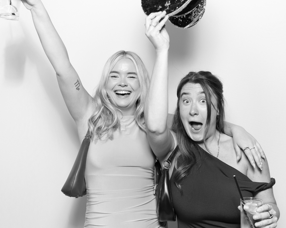 Two women celebrating with drinks and props, captured mid-laugh in a digital photo booth session.