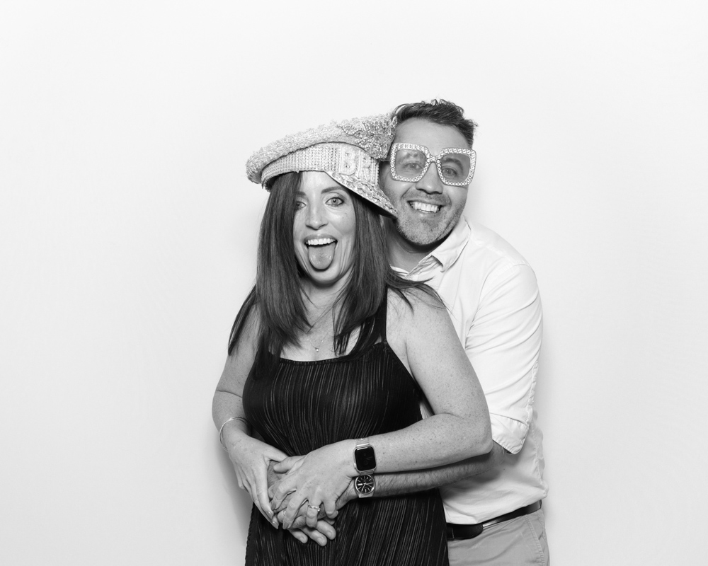 Couple posing with oversized party glasses and a "bride" hat, having fun in a black and white photo booth.