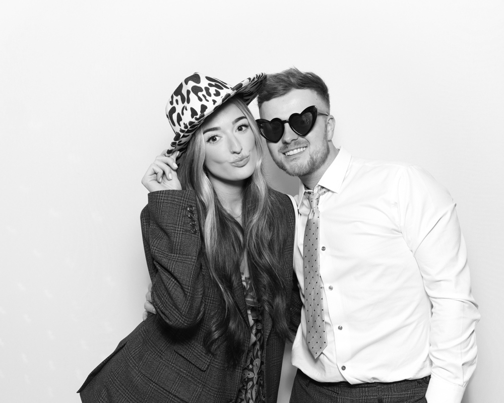Couple in party attire, posing with a leopard-print hat and heart sunglasses in a black and white booth photo.