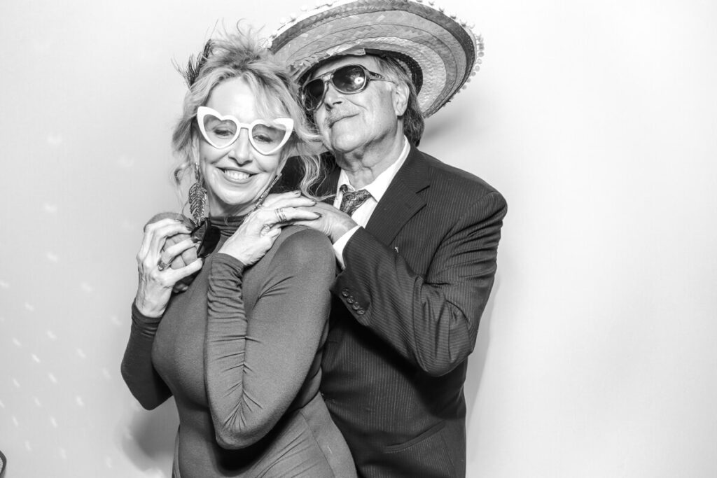Elegant older couple wearing fun props and laughing together in the black and white photo booth during a summer Kiftsgate Court Gardens wedding.