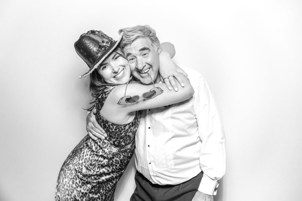 Female guest in a sequin hat hugging an older man and smiling in the black and white photo booth at a Kiftsgate Court Gardens wedding.