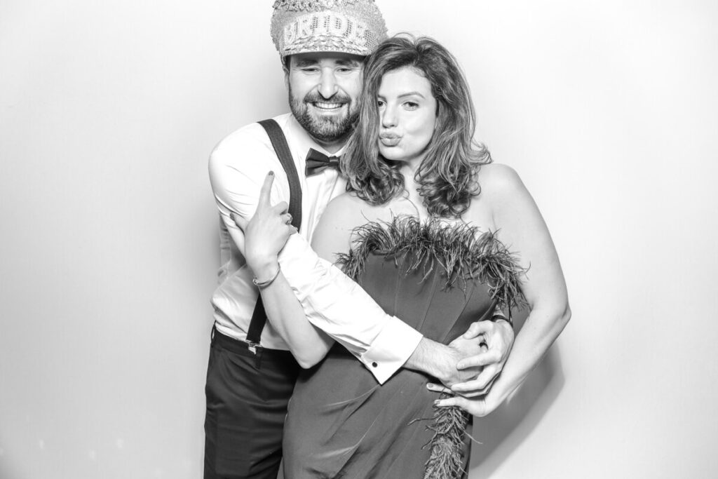 Bride and groom-style couple striking a pose in the black and white photo booth at a Kiftsgate Court Gardens wedding, with the groom wearing a "Bride" hat.