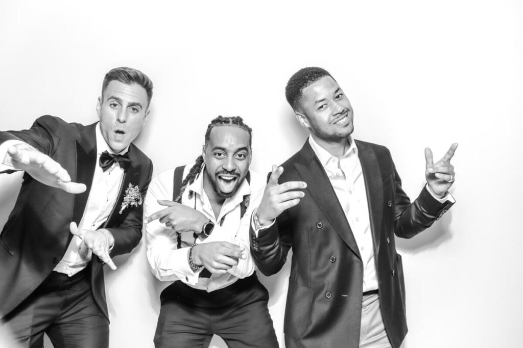 Three male guests in tuxedos and suits striking playful poses in the black and white photo booth during a chipping campden celebration.