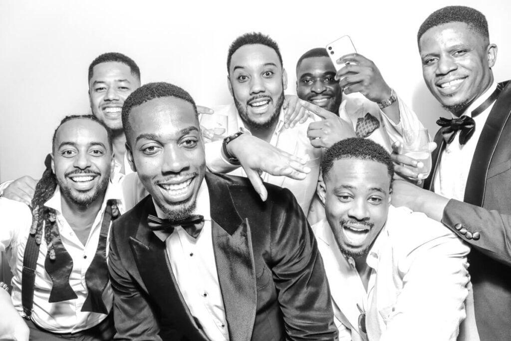 A group of groomsmen and guests smiling and laughing in the black and white photo booth at a wedding at Kiftsgate Court Gardens.