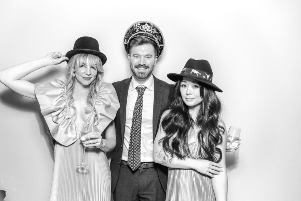 Three wedding guests wearing hats and holding drinks while posing in the black and white photo booth at Kiftsgate Court Gardens.