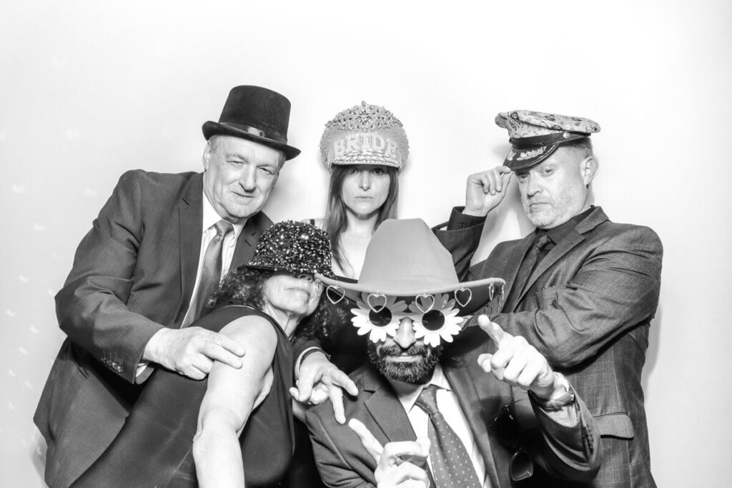 Wedding guests dressed in suits and party hats having fun with props in the black and white photo booth at a Kiftsgate Court Gardens marquee reception.