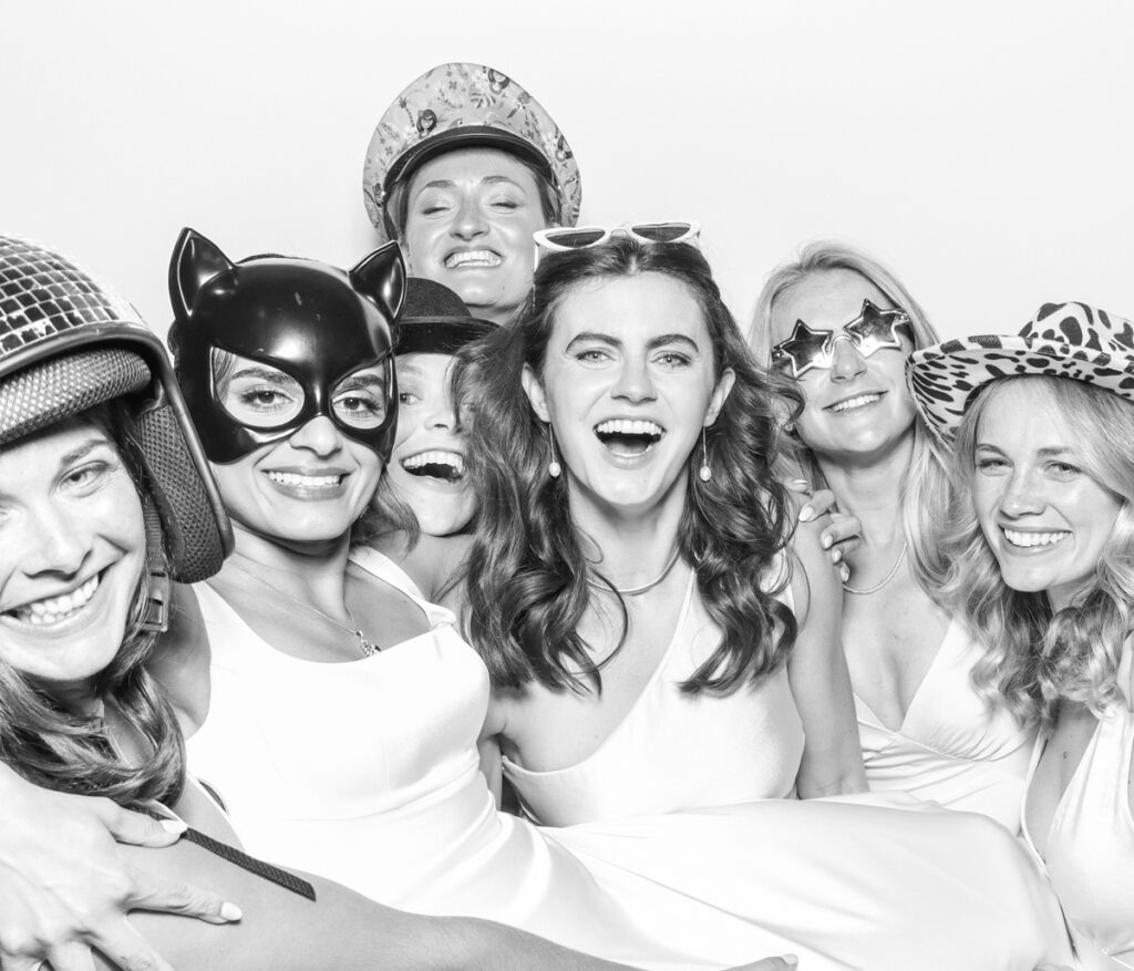 large group of wedding guests posing against a plain white backdrop, during the party entertainment in the evening, wearing fun party props and laughing for the camera, with black and white prints for the kardashian style photos